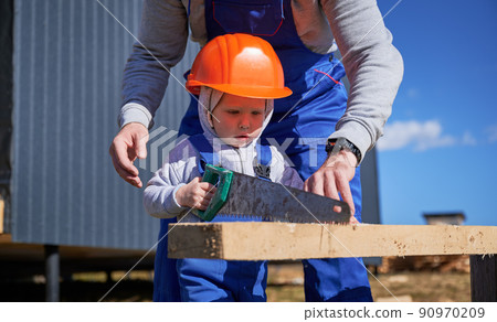 Father with toddler son building wooden frame house. Boy helping his dad, using hand saw to cut boards on construction site, wearing helmet and blue overalls on sunny day. Carpentry and family concept 90970209