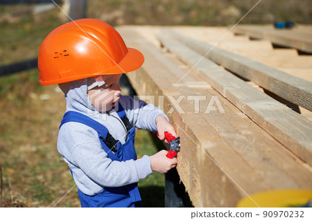 Boy toddler playing as builder on construction site. Child carpenter in orange helmet and blue overalls learning to build wooden frame house outdoor on sunny day. Carpentry and workshop concept. 90970232