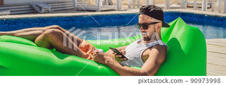 Young man enjoying leisure, lying on the air sofa lamzac, near the pool BANNER, LONG FORMAT 90973998