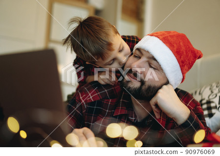 man i in santa hat with his little cute son laying with laptop on bed buying presents online 90976069