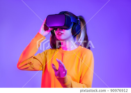 Young asian woman in orange sweatshirt wearing black vr headset watching playing and hand touching on the purple background. Metaverse concept vr future girl lifestyle 90976912