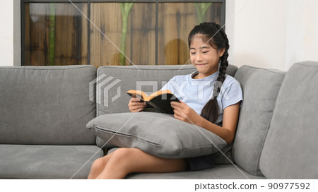 Smiling preteen girl relaxing on comfortable sofa and reading book 90977592