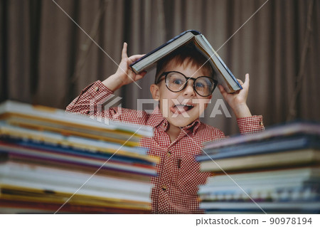 cute caucasian boy wearing glasses covering head with book. Cheerful boy peeking from behind piles of books 90978194