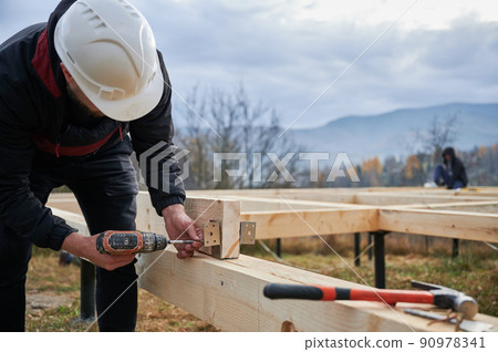 Man worker building wooden frame house on pile foundation. Carpenter installing furniture for wooden planks, using screwdriver. Carpentry concept. 90978341
