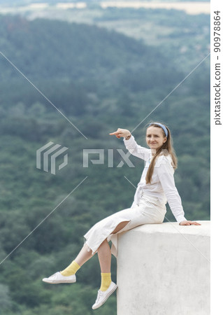 Young woman sits on balcony with her legs hanging down and points into the distance on mountains background. Vertical frame. Young woman sits on balcony with her legs hanging down and points into the distance on mountains background. Vertical frame. 90978864