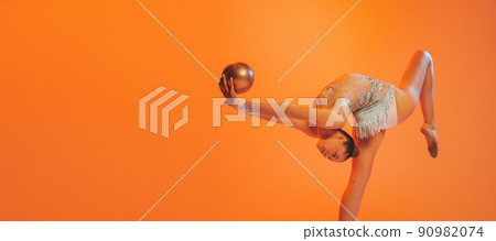 One professional rhythmic gymnastics artist training with golden color ball isolated on orange background. Concept of sport, action, aspiration, competition 90982074