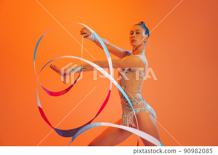 Young sportive girl, rhythmic gymnastics artist dancing isolated on orange color background. Concept of sport, action, aspiration, education, active lifestyle Young sportive girl, rhythmic gymnastics artist dancing isolated on orange color background. Concept of sport, action, aspiration, education, active lifestyle 90982085