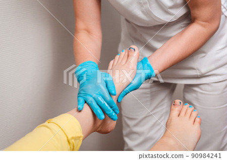 A podologist massages the client's feet. Close-up of legs and toenails with titanium thread and master's arms in medical gloves. The concept of chiropody and podology A podologist massages the client's feet. Close-up of legs and toenails with titanium thread and master's arms in medical gloves. The concept of chiropody and podology 90984241