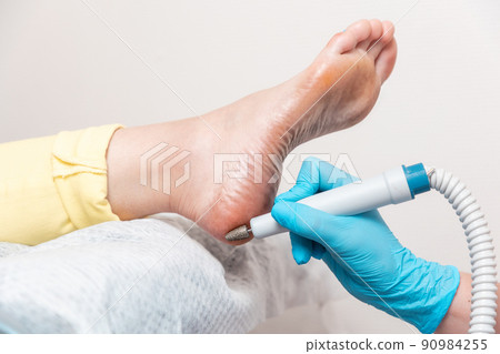 Pedicurist does hardware peeling of the skin of the heel. Close-up of dry foot and master's hand. The concept of professional podology and chiropody Pedicurist does hardware peeling of the skin of the heel. Close-up of dry foot and master's hand. The concept of professional podology and chiropody 90984255