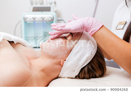 Cosmetologist's hands at protective gloves massages client's face with cosmetics mask. Side view. Concept of professional cosmetology Cosmetologist's hands at protective gloves massages client's face with cosmetics mask. Side view. Concept of professional cosmetology 90984268