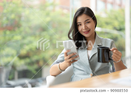 Smiling asian businesswoman using phone in office. Small business entrepreneur looking at her mobile phone and smiling. 90988550