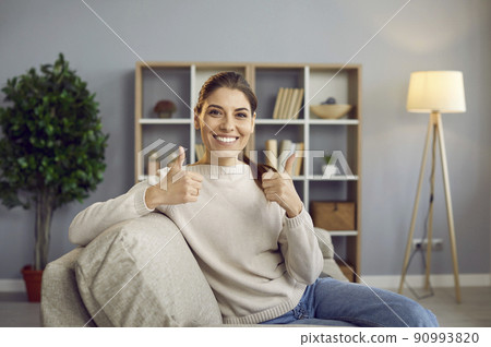 Happy young woman sitting on sofa at home, smiling and showing thumbs up gesture Happy young woman sitting on sofa at home, smiling and showing thumbs up gesture 90993820