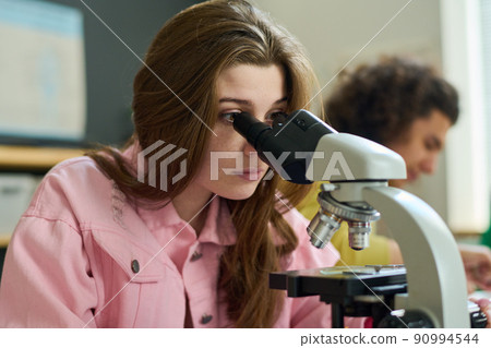 Serious teenage girl in pink jacket looking in microscope at chemistry lesson Serious teenage girl in pink jacket looking in microscope at chemistry lesson 90994544