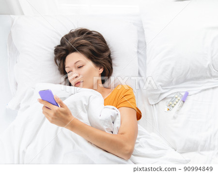 Top view on sick woman lying in bed with smartphone. Thermometer and pills on white bed linen. Internet surfing. 90994849