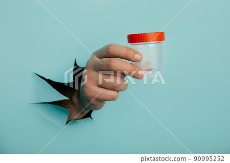 Male hand holds a plastic jar for urine tests, the hand is sticking out of a torn hole in a blue background 90995252