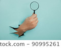 Male hand holds black magnifying glass in torn hole of blue background. Searching information concept 90995261