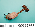 Hand holding a judge's gavel through torn blue paper wall. Law and courts, triumph of justice 90995263