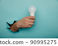 Hand holding an incandescent led light bulb from a torn hole in blue paper 90995275