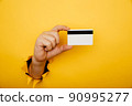 Male hand is holding credit bank card in torn hole of yellow background. Online shopping, purchasing products concept. 90995277