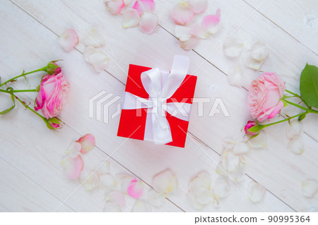 On a white wooden background there are roses and petals forming a circle, inside there is a gift in a red box and a white ribbon. High quality photo 90995364