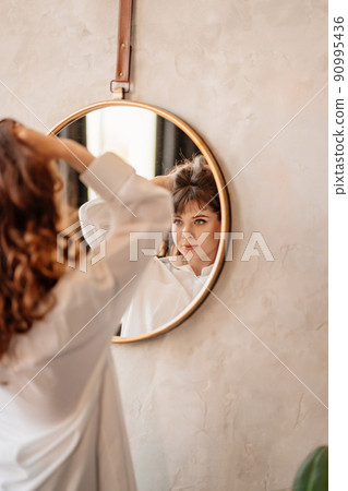 a beautiful woman in a white shirt looks at her reflection in a round mirror. a beautiful woman in a white shirt looks at her reflection in a round mirror. 90995436