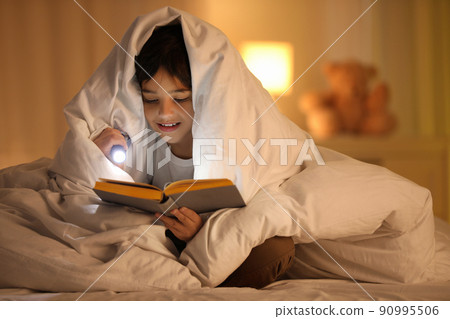 Boy with flashlight reading book under blanket at home 90995506