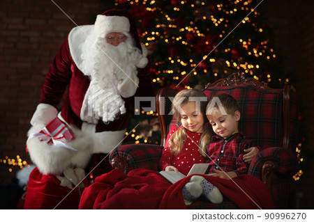 Santa Claus sneaking in with gifts while children reading book near Christmas tree indoors Santa Claus sneaking in with gifts while children reading book near Christmas tree indoors 90996270