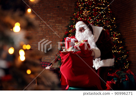 Santa Claus packing gifts into bag near Christmas tree Santa Claus packing gifts into bag near Christmas tree 90996271