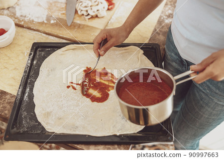 A woman spreads tomato sauce on pizza dough on a baking dish 90997663