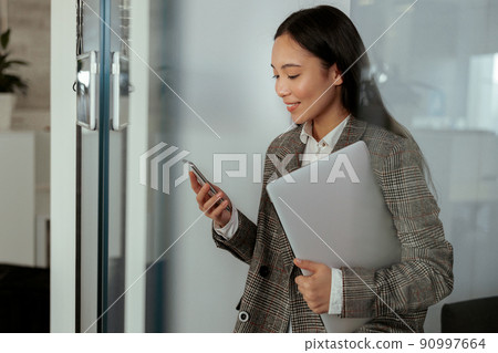 Asian business woman with a smile standing holding laptop and mobile phone at the office Asian business woman with a smile standing holding laptop and mobile phone at the office 90997664