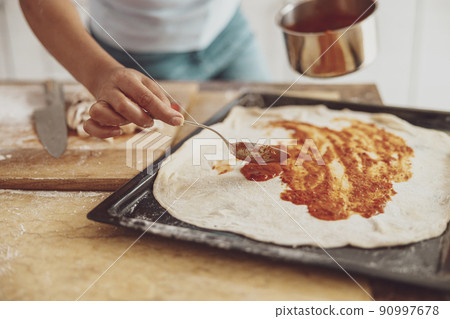 A woman's hand pours tomato sauce over the surface of a pizza laid out on the baking tray. A woman's hand pours tomato sauce over the surface of a pizza laid out on the baking tray. 90997678