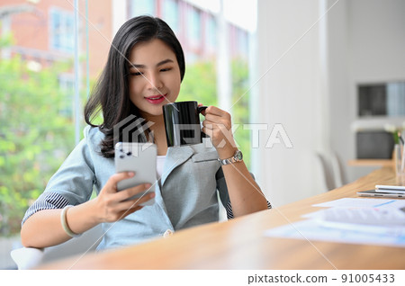 Gorgeous Asian businesswoman sipping coffee and reading a business news on her phone 91005433