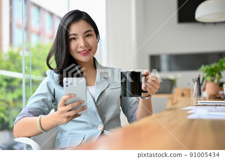 Charming young asian businesswoman sipping coffee while using a smartphone 91005434