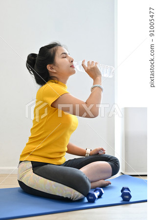 Exhausted young asian woman drinking water from the bottle on the yoga mat. 91005477