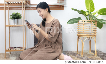 Young Asian woman happily using her mobile phone to chat or talking with her friends, Entertaining or relaxing using mobile phone technology, Stay at home for long weekend, Happy lifestyle. 91006844