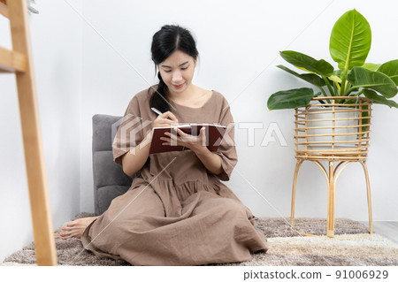 Asian woman sits in her living room writing a diary or notebook on the carpet, Weekends or holidays at home, Relaxation, Happy time, Living at home, Sit comfortably on vacation, Comfortable corner. 91006929