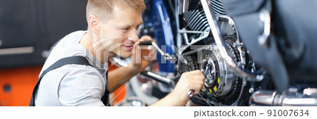 Young male locksmith disassembles motorcycle engine on bench in garage Young male locksmith disassembles motorcycle engine on bench in garage 91007634