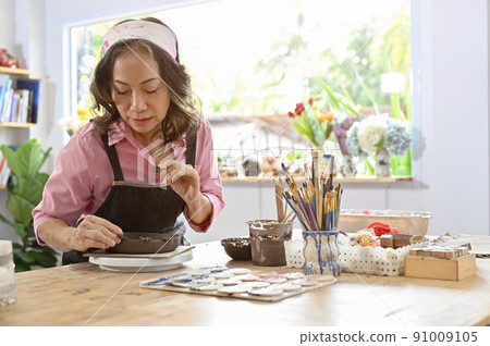 Professional asian aged woman decorating her plate with clay accessories on the table. 91009105