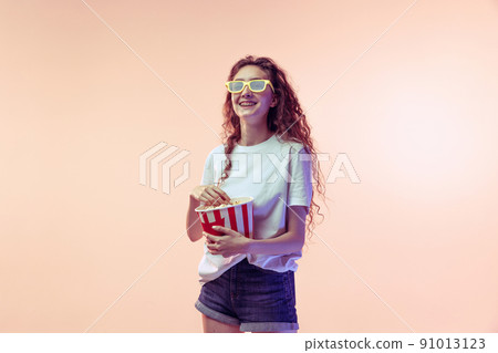 Portrait of young girl in casual outfit posing with popcorn basket isolated over pink studio background in neon light 91013123