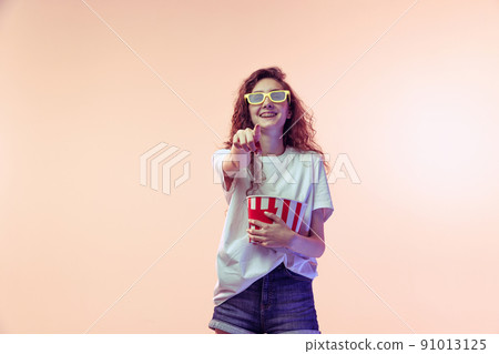 Portrait of young girl in casual outfit posing with popcorn basket isolated over pink studio background in neon light. Comedy film Portrait of young girl in casual outfit posing with popcorn basket isolated over pink studio background in neon light. Comedy film 91013125