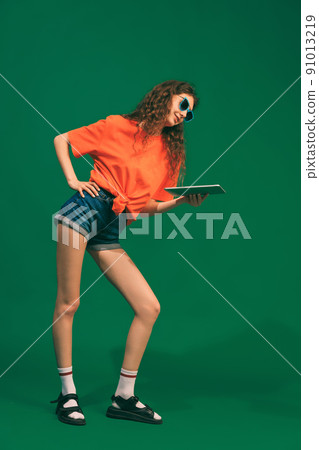 Portrait of young girl in casual outfit, shorts and T-shirt, holding tablet, posing isolated over green studio background. Online services 91013219