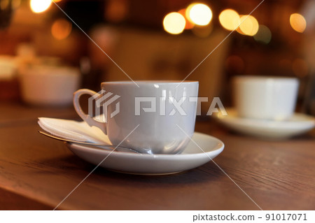 White tea cup standing on saucer in soft focus on naturally blurred background with bokeh lights. Coffee cup on rustic wooden table. Coffee, tea house. The concept of a cozy pastime, tea ceremony 91017071