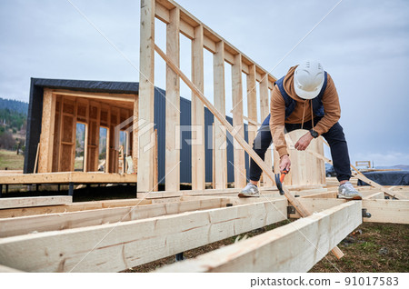 Man worker building wooden frame house on pile foundation. Carpenter hammering nail into wooden joist, using hammer. Carpentry concept. Man worker building wooden frame house on pile foundation. Carpenter hammering nail into wooden joist, using hammer. Carpentry concept. 91017583