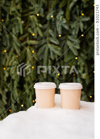 Two paper coffee cups outdoors in the snow against the backdrop of a beautiful green nobilis wall, coffee to go concept. Walks in the open air. Two paper coffee cups outdoors in the snow against the backdrop of a beautiful green nobilis wall, coffee to go concept. Walks in the open air. 91021743