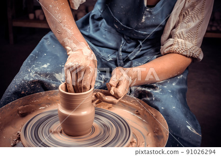 Close-up A woman potter in beautifully sculpts a deep bowl of brown clay and cuts off excess clay on a potter's wheel in a beautiful workshop Close-up A woman potter in beautifully sculpts a deep bowl of brown clay and cuts off excess clay on a potter's wheel in a beautiful workshop 91026294