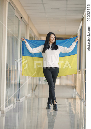 Woman with ukrainian flag posing for a photo. Young pretty girl standing in front of glass wall. Brunette woman wearing black clothes and support Ukraine. 91032536