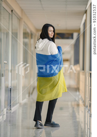 Woman with ukrainian flag posing for a photo. Young pretty girl standing in front of glass wall. Brunette woman wearing black clothes and support Ukraine. 91032537