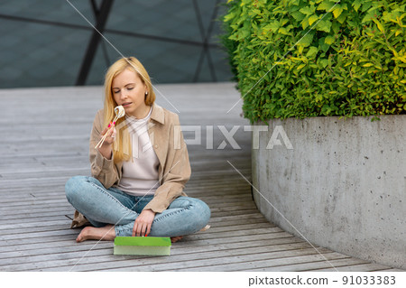 Young beautiful blond woman eating sushi outdoors, on the wooden terrace, by modern building in the city. Tasty food to go. Girl has lunch break, spending time outside and eating Asian food. City life Young beautiful blond woman eating sushi outdoors, on the wooden terrace, by modern building in the city. Tasty food to go. Girl has lunch break, spending time outside and eating Asian food. City life 91033383