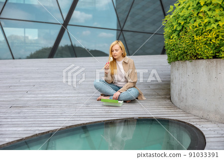 Young beautiful blond woman eating sushi outdoors, on the wooden terrace, by modern building in the city. Tasty food to go. Girl has lunch break, spending time outside and eating Asian food. City life 91033391