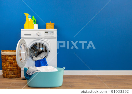 Washing machine with laundry on blue wall background Washing machine with laundry on blue wall background 91033392